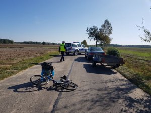 na pierwszym planie widać policjanta, ponadto na środku jezdni  leży rower  , z prawej strony stoi samochód z przyczepką  ,w poprzek jezdni ustawiony jest radiowóz policyjny