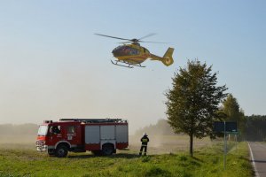 Lądujący na polu śmigłowiec Lotniczego Pogotowia Ratunkowego. Po lewej stronie wóz strażacki oraz strażacy zabezpieczający lądowanie.