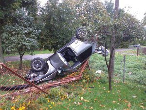 Rozbity pojazd marki Audi A3 wywrócony na dach Obok zniszczone metalowe ogrodzenie posesji.