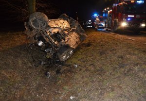 Rozbity pojazd marki Mercedes leżący na dachu w przydrożnym rowie. Po prawej na jezdni zaparkowane wozy strażackie z włączonymi światłami