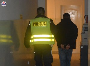 zatrzymany ubrany w czarny kaszkiet, czarną kurkę i jeansowe spodnie prowadzony przez policjanta w kamizelce odblaskowej