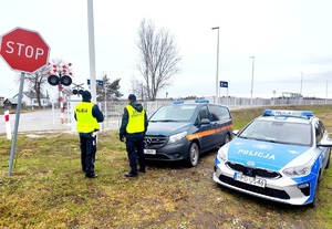 umundurowany policjant oraz strażnik ochrony kolei w rejonie przejazdu kolejowego, w tle oznakowane pojazdy służbowe