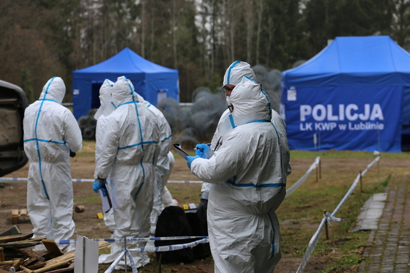 osoby ubrane w białe kombinezony stojące obok niebieskiego namiotu z napisem policja